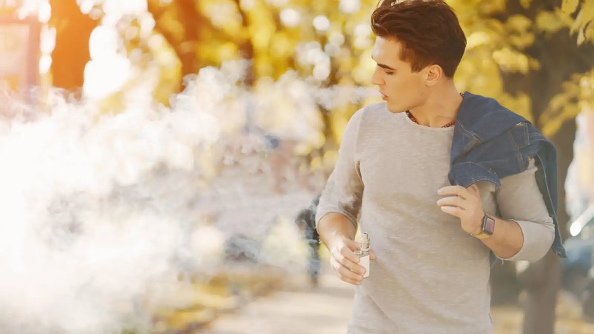 Jeune homme marchant dans un parc avec une cigarette électronique dans la main et soufflant la vapeur